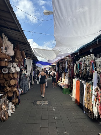 A vibrant, bustling market aisle is lined with stalls featuring a variety of colorful clothing, woven baskets, and textiles. Shoppers walk through the narrow path, shaded by large white tarps overhead. The ambiance suggests a lively, open-air environment.