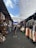 A vibrant, bustling market aisle is lined with stalls featuring a variety of colorful clothing, woven baskets, and textiles. Shoppers walk through the narrow path, shaded by large white tarps overhead. The ambiance suggests a lively, open-air environment.