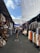 A vibrant, bustling market aisle is lined with stalls featuring a variety of colorful clothing, woven baskets, and textiles. Shoppers walk through the narrow path, shaded by large white tarps overhead. The ambiance suggests a lively, open-air environment.