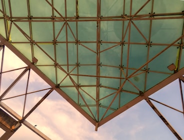 A triangular glass roof structure with metal support beams, featuring a grid pattern. The sky is visible in the background, with a small bird perched on one of the beams.