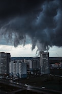 Dark, swirling tornado-like clouds hover ominously over a cityscape with tall residential buildings. The atmosphere is tense as the storm threatens the urban area below. The scene is set in a late afternoon or early evening, casting a dramatic and shadowy effect over the landscape.