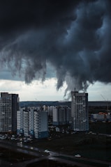 Dark, swirling tornado-like clouds hover ominously over a cityscape with tall residential buildings. The atmosphere is tense as the storm threatens the urban area below. The scene is set in a late afternoon or early evening, casting a dramatic and shadowy effect over the landscape.