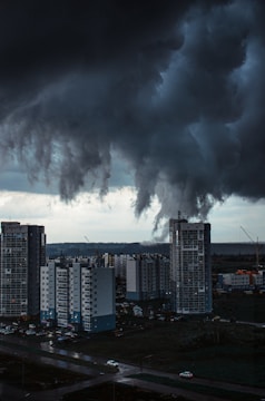 Dark, swirling tornado-like clouds hover ominously over a cityscape with tall residential buildings. The atmosphere is tense as the storm threatens the urban area below. The scene is set in a late afternoon or early evening, casting a dramatic and shadowy effect over the landscape.