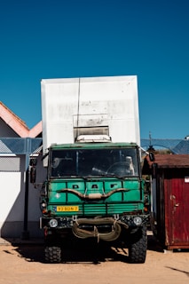 A large green truck with a box-like structure on its back, parked beside a white wall and a smaller wooden building. The truck has a worn appearance and displays a license plate. The sky is clear and blue.