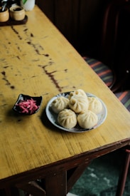 A wooden table with a rustic appearance holds a plate of steamed buns and a small dish of pickled vegetables. The background features a wooden chair with colorful, striped fabric.