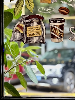 Illustrated bags of ground dark roasted coffee labeled as organic are depicted alongside a takeaway coffee cup and a coffee mug with steam above, placed against a backdrop of greenery and a city street viewed through a window.