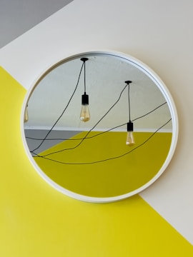 A round mirror with a white frame reflects hanging Edison-style light bulbs and black wires on a textured ceiling. The mirror is mounted on a wall painted in geometric patterns of yellow and white, creating a modern and abstract appearance.