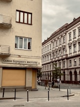 An urban street scene featuring two buildings with classic architectural designs. The building on the left has a beige facade and displays a sign written in Ukrainian, while the building on the right shows ornate detailing with arched windows. Pedestrians are visible walking past the buildings, and there are a few young trees lining the sidewalk.