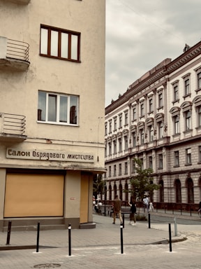 An urban street scene featuring two buildings with classic architectural designs. The building on the left has a beige facade and displays a sign written in Ukrainian, while the building on the right shows ornate detailing with arched windows. Pedestrians are visible walking past the buildings, and there are a few young trees lining the sidewalk.