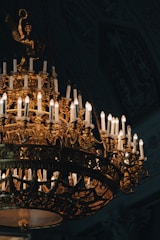An ornate, antique chandelier decorated with numerous lit candles casting a warm glow. The intricate design features gold detailing and sculptural elements, creating an opulent and elegant appearance. The background is dark, highlighting the splendor of the chandelier.