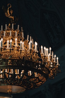 An ornate, antique chandelier decorated with numerous lit candles casting a warm glow. The intricate design features gold detailing and sculptural elements, creating an opulent and elegant appearance. The background is dark, highlighting the splendor of the chandelier.