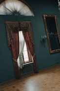 A room featuring an elegant window with heavy, red and gold drapes. The window is situated on a dark teal wall, beneath an ornate half-moon fanlight. Beside the window is a large mirror with an intricate gold frame. The floor is made of polished wood with an inlaid pattern. Wall sconces with candle-style lights add to the classical decor.