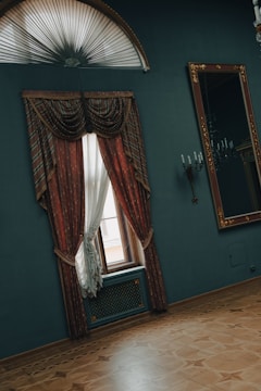 A room featuring an elegant window with heavy, red and gold drapes. The window is situated on a dark teal wall, beneath an ornate half-moon fanlight. Beside the window is a large mirror with an intricate gold frame. The floor is made of polished wood with an inlaid pattern. Wall sconces with candle-style lights add to the classical decor.