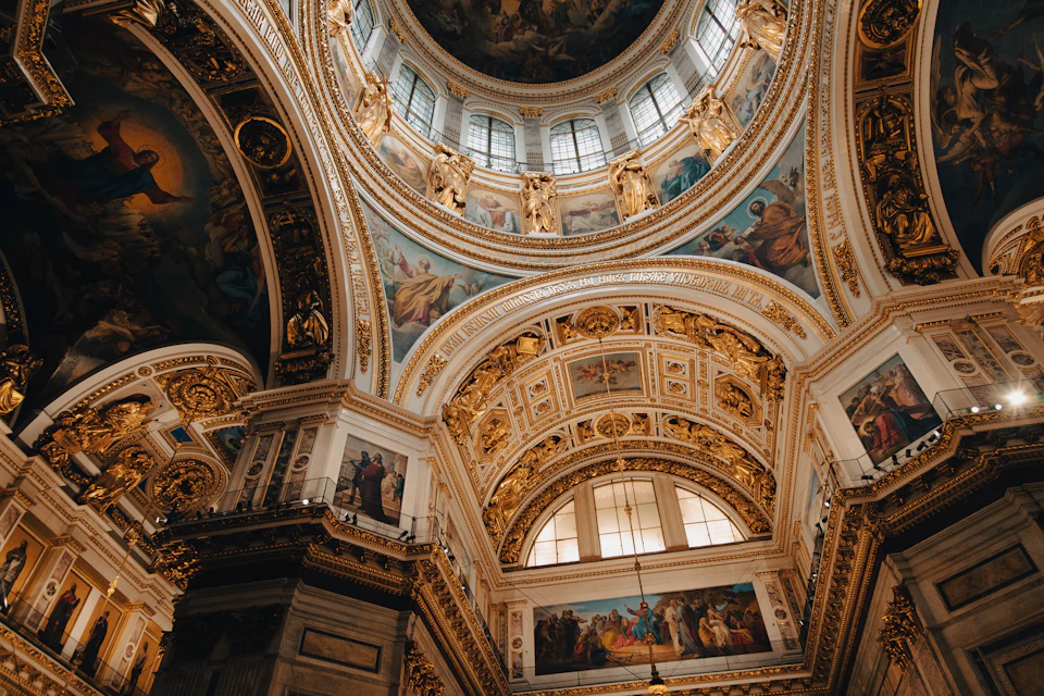 The image depicts a grand, ornate room with a large dome at its center. The dome is adorned with intricate gold and white designs that shimmer under the light. The walls surrounding the dome are also embellished with gold and white patterns, creating a harmonious blend of colors throughout the space. The ceiling features a series of windows that allow natural light to filter in, enhancing the room's overall ambiance. The perspective of the image is from below, looking up at the dome, which emphasizes its imposing presence within the room.