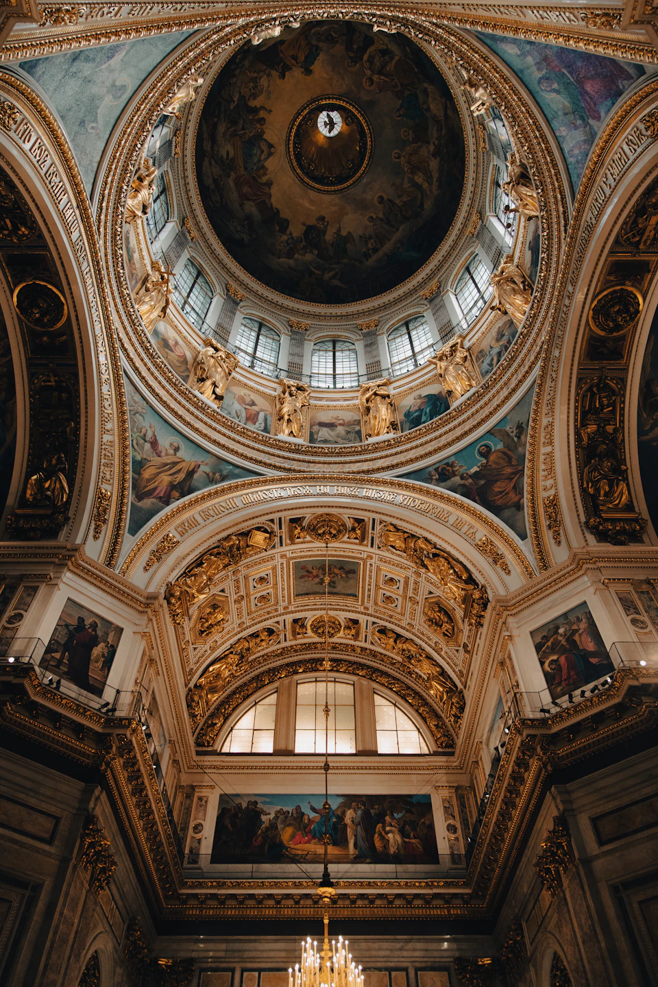 The image depicts a grand, ornate room with a large dome at its center. The dome is adorned with intricate gold and blue designs that shimmer under the light. The ceiling features numerous windows, allowing natural light to filter into the space. The walls are embellished with paintings depicting various scenes of people and animals. A chandelier hangs from the ceiling, casting a warm glow throughout the room.