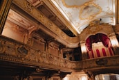 A lavishly decorated interior of a theater or opera house with ornate gold detailing, red velvet curtains, and a painted ceiling. The architectural style is baroque, featuring elaborate moldings and carvings, with dramatic lighting accentuating the opulence.