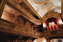 A lavishly decorated interior of a theater or opera house with ornate gold detailing, red velvet curtains, and a painted ceiling. The architectural style is baroque, featuring elaborate moldings and carvings, with dramatic lighting accentuating the opulence.