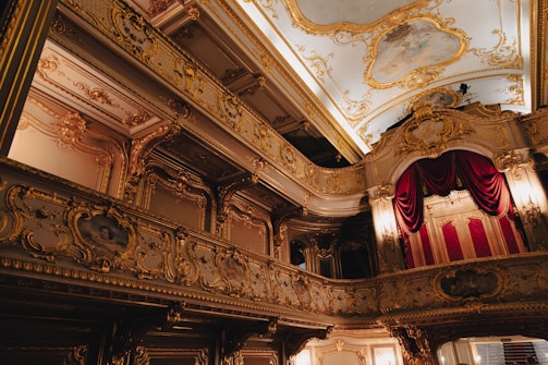 A lavishly decorated interior of a theater or opera house with ornate gold detailing, red velvet curtains, and a painted ceiling. The architectural style is baroque, featuring elaborate moldings and carvings, with dramatic lighting accentuating the opulence.