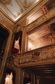 An opulent and ornately decorated interior space with elaborate gold detailing and moldings. Rich red velvet curtains complement the luxurious setting and intricate carvings adorn the walls and ceilings. The warm lighting enhances the grandeur of the space.