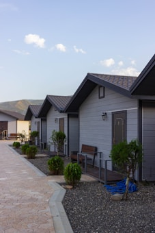 A row of small, single-story cabins with dark roofs and light gray siding. Each cabin has a front door and a few small shrubs or bushes planted in front. The cabins are aligned along a paved stone walkway with a clear blue sky and distant mountains visible in the background.