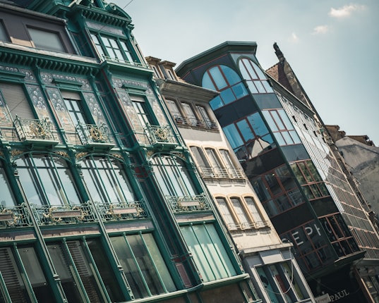 A row of architecturally diverse buildings featuring an Art Nouveau style with intricate floral designs, ornate railings, and large windows reflecting the sky. The structures vary in color and material, showcasing both historical and modern elements.