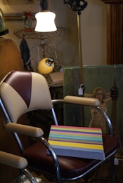 A vintage-style room with a mix of old and contemporary elements featuring a retro chair with cream and brown upholstery. On the chair rests a canvas with multicolored horizontal stripes. The background includes an impressionistic painting of a woman, various antique objects, a tall floor lamp with an ornate black stand, and a hanging white lamp illuminating the scene.