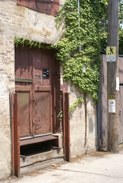 A rusty, metal door set within a weathered brick wall, partially covered by green ivy. The door has a sign saying 'Ring Bell'. Next to the door is a utility pole with various notices attached, including a speed limit sign. The pavement is cracked and uneven, adding to the aged appearance of the scene.