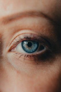 A close-up view of a human eye with a blue iris, surrounded by skin showing fine details and natural textures. Light reflects off the eye, highlighting the intricate patterns in the iris.