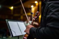 A person in a dark suit is holding a violin and bow in a dimly lit room. The focus is on the hand, the violin's scroll, and a music stand with sheet music nearby. Warm lighting is present, creating a cozy ambiance.