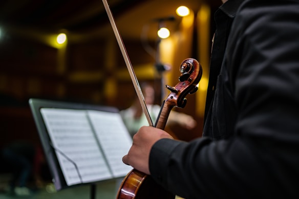 A person in a dark suit is holding a violin and bow in a dimly lit room. The focus is on the hand, the violin's scroll, and a music stand with sheet music nearby. Warm lighting is present, creating a cozy ambiance.