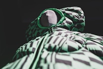 A mannequin displays a puffy jacket with a striking green and black geometric pattern, featuring a hood and zippered front. The dramatic lighting highlights the texture and design, creating a bold and modern aesthetic.
