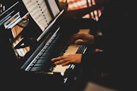 Hands are playing the keys of a piano with a music book open in front of them. The lighting creates a warm and intimate atmosphere.