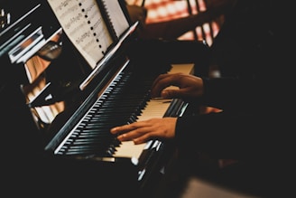 Hands are playing the keys of a piano with a music book open in front of them. The lighting creates a warm and intimate atmosphere.