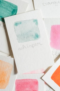 Several small watercolor cards are scattered, each featuring a different color wash. The prominent card displays a light teal watercolor with the word 'whisper' written underneath. Other visible colors include pink, orange, and dark teal.