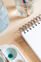 A close-up of art supplies on a wooden table, including a spiral-bound notebook, a brush in a watercolor palette with blue paint, and two clear jars filled with water and paintbrushes.