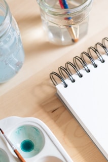 A close-up of art supplies on a wooden table, including a spiral-bound notebook, a brush in a watercolor palette with blue paint, and two clear jars filled with water and paintbrushes.
