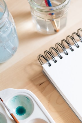 A close-up of art supplies on a wooden table, including a spiral-bound notebook, a brush in a watercolor palette with blue paint, and two clear jars filled with water and paintbrushes.