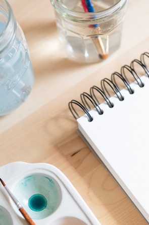 A close-up of art supplies on a wooden table, including a spiral-bound notebook, a brush in a watercolor palette with blue paint, and two clear jars filled with water and paintbrushes.