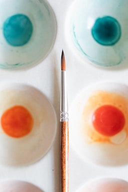 A watercolor palette with various paint wells filled with shades of blue and orange, coupled with a fine-tipped paintbrush placed diagonally across the palette. The palette is white, and the paint wells show a blend of colors demonstrating artistic use.