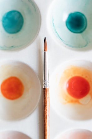 A watercolor palette with various paint wells filled with shades of blue and orange, coupled with a fine-tipped paintbrush placed diagonally across the palette. The palette is white, and the paint wells show a blend of colors demonstrating artistic use.