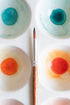 A watercolor palette with various paint wells filled with shades of blue and orange, coupled with a fine-tipped paintbrush placed diagonally across the palette. The palette is white, and the paint wells show a blend of colors demonstrating artistic use.