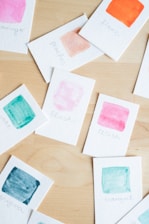 Several small cards are scattered on a wooden surface, each with a watercolor paint swatch and handwritten label. The colors include pink, orange, teal, and peach.