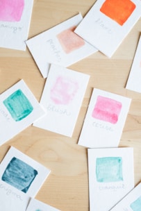 Several small cards are scattered on a wooden surface, each with a watercolor paint swatch and handwritten label. The colors include pink, orange, teal, and peach.