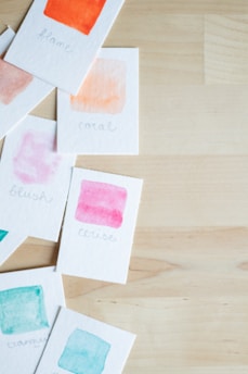 Multiple paint swatches on cards scattered over a light wooden surface, featuring various watercolor shades with handwritten labels, including 'flame', 'coral', 'blush', 'cerise', and 'turquoise'.