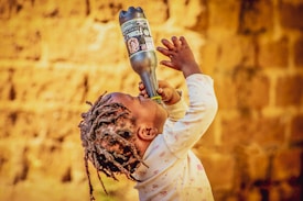 A young child with braided hair is drinking from a large bottle while looking upwards, against a backdrop of a textured, warm-colored wall. The child is wearing a long-sleeved shirt with a subtle pattern.