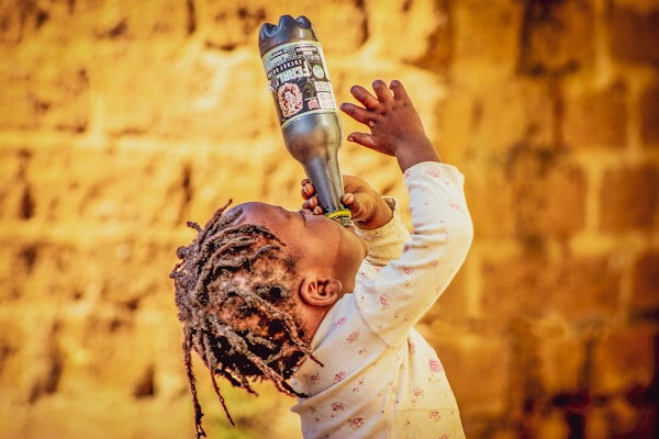 A young child with braided hair is drinking from a large bottle while looking upwards, against a backdrop of a textured, warm-colored wall. The child is wearing a long-sleeved shirt with a subtle pattern.