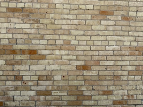 A wall composed of light brown and beige bricks arranged in a uniform pattern. Each brick has a slightly weathered texture, contributing to an overall rustic appearance. The mortar between the bricks is gray and evenly applied.