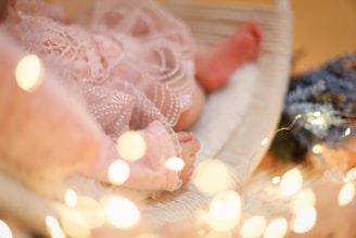 A close-up of a baby's feet and hands nestled in delicate lace fabric. The scene is warmly illuminated with soft, glowing fairy lights. The baby appears to be resting on a cozy surface, with a backdrop of blue-toned flowers adding a serene touch.