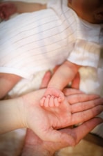 A tender moment is captured as an adult hand gently cradles the small hand of a baby, showcasing the contrast between their sizes. The baby is wearing a white outfit with thin black stripes, and the setting exudes warmth and intimacy.