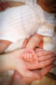 A tender moment is captured as an adult hand gently cradles the small hand of a baby, showcasing the contrast between their sizes. The baby is wearing a white outfit with thin black stripes, and the setting exudes warmth and intimacy.